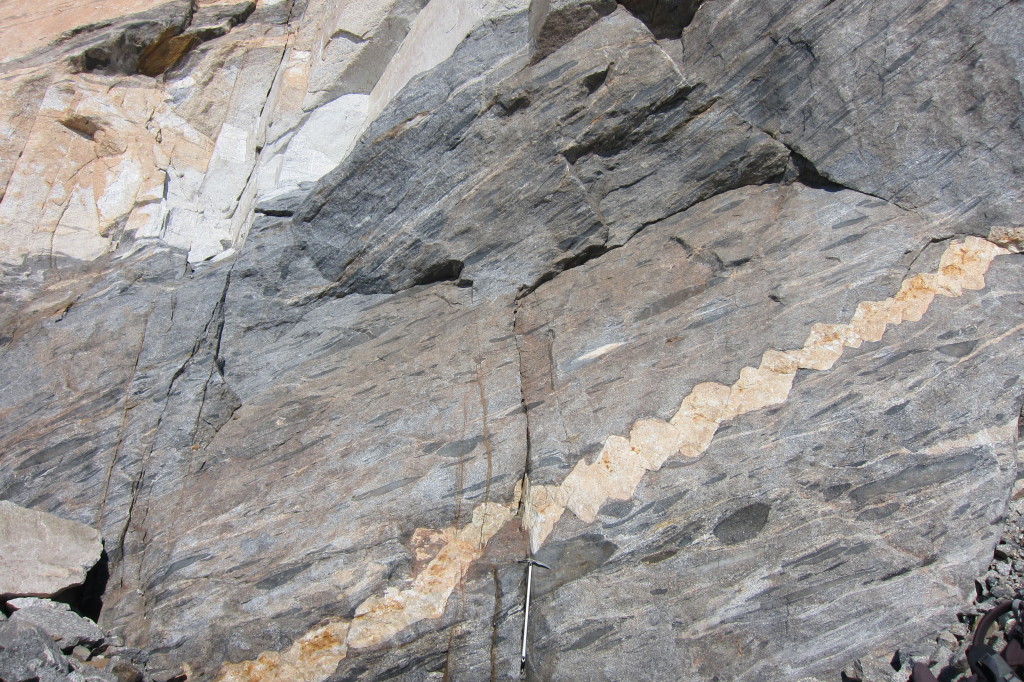 Figure 13. A dark grey orthogneiss containing strained mafic enclaves is cross-cut by a leucogranite dike that shows domino boudinage. Ice axe for scale.