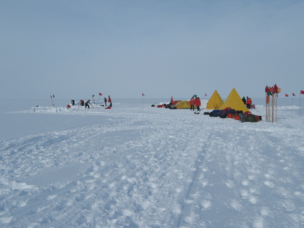 Figure 4. Several participants at the “Happy Camper” campsite dig snow trenches, while others set up tents. Red and green flags mark safe routes in case of poor-visibility conditions.