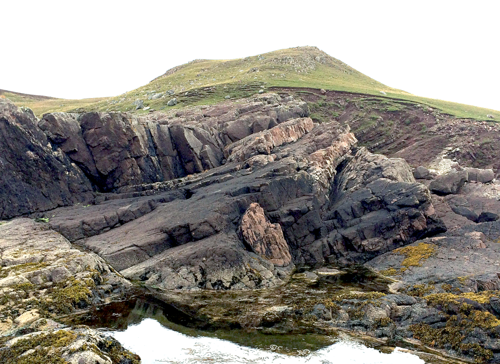 Fig. 3 – The Stac Fada Member fills most of this photo, including the ~5 metre tall subvertical faces to the left. The paler brown rocks are layers of pre-existing sandstone that were disrupted and folded by the fast-moving ejecta blanket. The nose of one of these folds is clearly visible in the centre of the photograph.