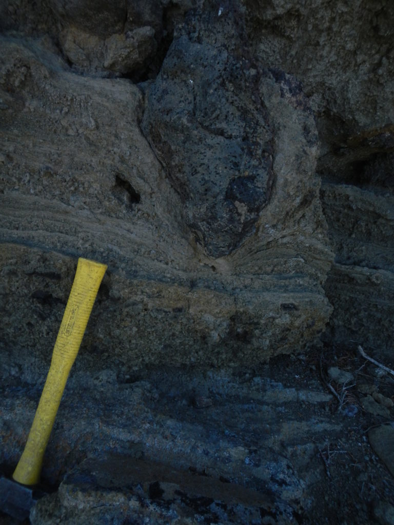 A hydrovolcanic feature in the Reed Rock deposits known as a 'bomb sag'. The lava bomb in the upper portion of the photo was deposited into the lower layers when they were saturated with water. The layers were thus plastically deformed. Photo by Matthew Nikitczuk.